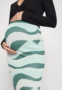 Pletená těhotenská sukně se zelenými a bílými vlnitými vzory, přiléhavý střih a měkká textura, doplněná černým topem s dlouhým rukávem.