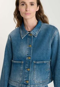 Kvinna med långt brunt hår som bär en knappad blå denimjacka med stora framfickor mot en enkel bakgrund.