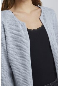 Ljusblå, stickad cardigan med ett strukturerat mönster, öppen front och ribbade sömmar, bärs över en svart linne med spetskant.