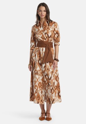 Kvinde iført en knapoplukkelig, brun- og hvidmønstret midi-kjole med trekvartlange ærmer, et bredt brunt bælte og brune T-rem sko.