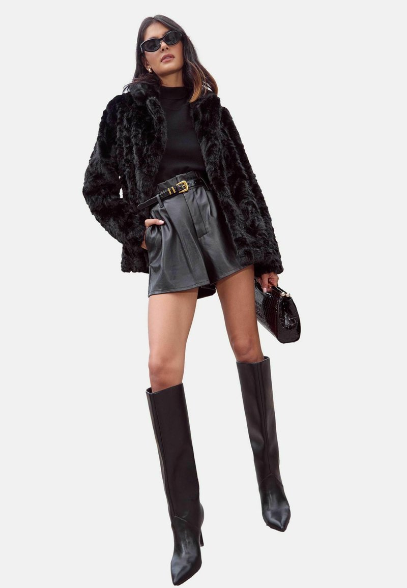 Melnas mākslīgās kažokādas mēteles pāri melnam tērpam, apvienota ar augsto jostasvietu ādas šortiem, ceļgalu augstiem melniem zābakiem un melnu rokassomu.