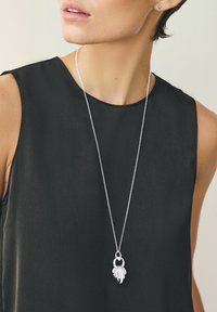 Zilveren hangersnoer met een ketting en een ingewikkeld ontworpen bedel die lijkt op gelaagde bladeren. Gedragen over een zwart top.
