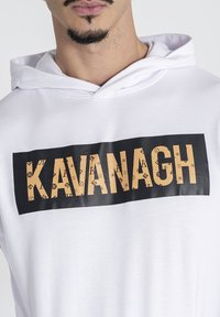 Biela mikina s kapucňou s čiernym obdĺžnikovým pásom, na ktorom je zlatým písmom napísané slovo "KAVANAGH", doplnené rôznymi logami a symbolmi okolo písmen.