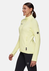 Giacca in pile giallo chiaro con colletto alto, cerniera longitudinale e logo nero sulla manica. Presenta tasche frontali con cerniera e maniche lunghe.