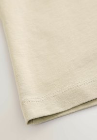 Tissu beige avec une texture fine en tricot et un ourlet soigneusement cousu plié au bord, présenté sur un fond clair.