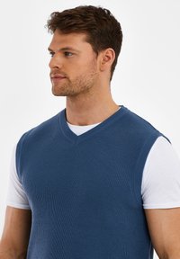 Blauwe geribbelde mouwloze trui met V-hals, gedragen over een wit t-shirt. Heeft een gestructureerde stof en een casual pasvorm.