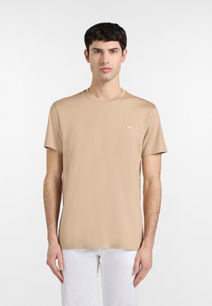 Giovane uomo con barba corta e ricci scuri, indossa una maglietta beige a maniche corte senza decorazioni e pantaloni bianchi, in piedi su uno sfondo bianco.