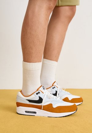 Nogi w beżowych szortach, białych prążkowanych skarpetach za kostkę i białych butach Nike Air Max z pomarańczowymi zamszowymi akcentami i czarnymi logo Swoosh.
