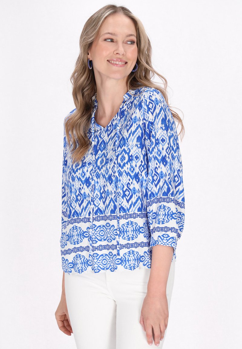 Blusa blu a fantasia con scollatura a V, maniche lunghe e dettagli bianchi. Realizzata in tessuto leggero con disegni geometrici intricati.