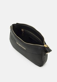 Musta nahkakäsilaukku, jossa on teksturoitu pinta, kultainen vetoketju ja logo-yksityiskohtia. Auki oleva yläosa paljastaa tilavan sisätilan ja pienen taskun.