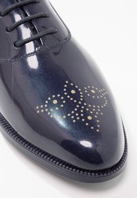 Chaussure habillée glossy bleu marine avec un motif complexe de pois clairs sur le bout et des lacets noirs sur fond blanc.