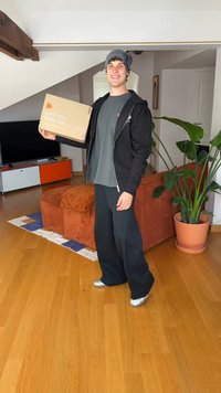 Caixa com texto "Ame-me. Use-me." segurada por uma pessoa com um capuz preto e camisa cinza, usando um gorro, calças largas pretas e sapatos prateados.