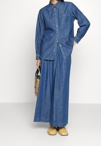 Nainen, joka pukeutuu löysään farkkupaitaan ja leveälahkeisiin farkkuihin, pitää kädessään punottua käsilaukkua ja on jalassaan vaaleanruskeat mokkasiinityyliset kengät.