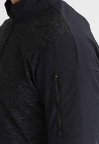 Primer plano de una chaqueta deportiva negra con tejido texturizado en el torso y tela suave en la manga, que cuenta con un bolsillo vertical con cremallera.