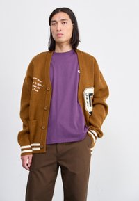 Bruine gebreide cardigan met witte strepen op de mouwen, voorzien van vo pockets en grafisch detail. Gecombineerd met een paarse T-shirt en bruine broek.