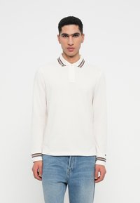 Polo de mangas longas branco com colarinho e punhos às riscas. Apresenta um bordado discreto de logotipo no peito. Usado com jeans azul claro.