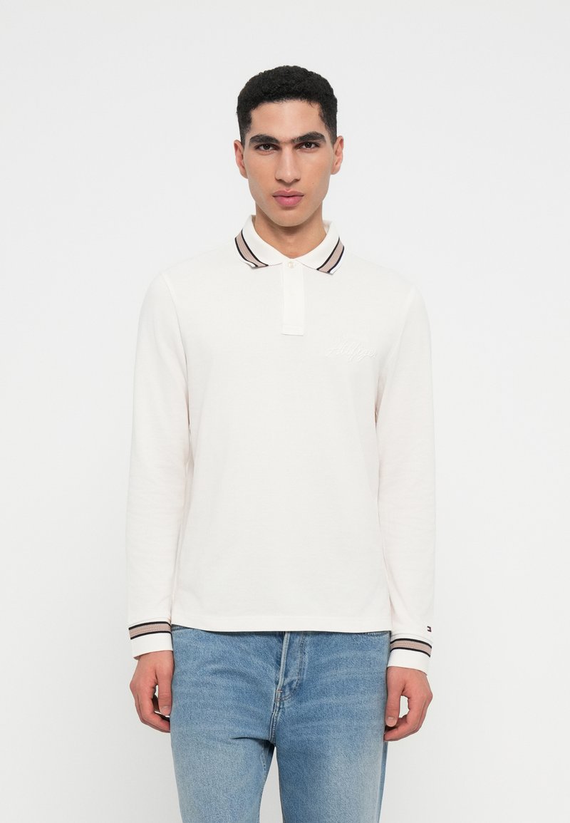 Polo de mangas longas branco com colarinho e punhos às riscas. Apresenta um bordado discreto de logotipo no peito. Usado com jeans azul claro.