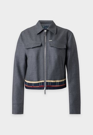 Jeune femme aux longs cheveux foncés portant une veste grise, un body noir, un jean foncé et des sandales noires, debout les mains dans les poches.