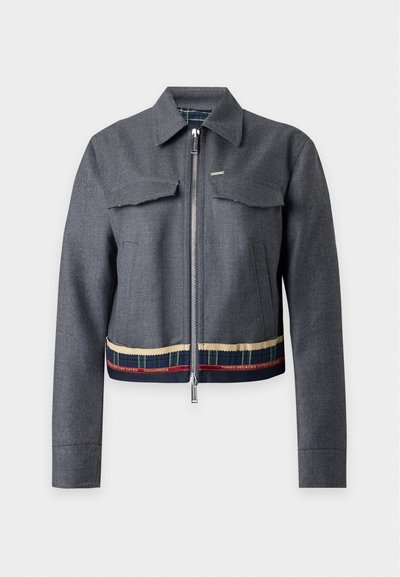 Veste grise zippée avec col, deux poches poitrine à rabat, et bordure rayée décorative avec texte en bas de l’ourlet sur fond blanc.