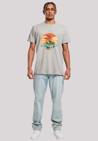 Graues Baumwoll-T-Shirt mit einem runden Grafikmuster von Bergen und Bäumen in Orange, Grün und Blau, kombiniert mit hellblauen Jeans und weißen Sneakers.