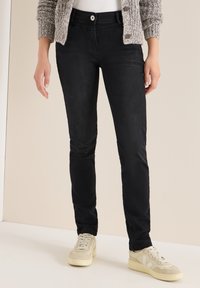 Jeans negros ajustados con una textura suave, que presentan un cierre de botón frontal. Combinados con zapatillas de color beige claro y un cárdigan de punto.