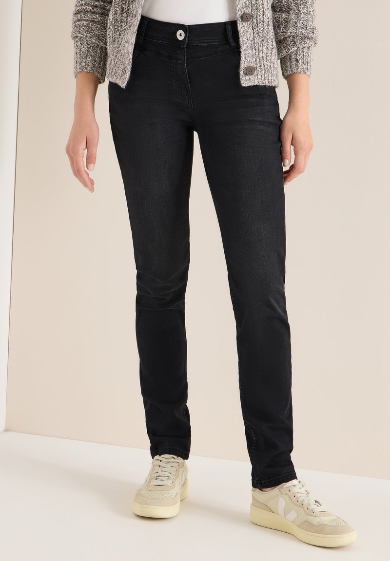 Jeans negros ajustados con una textura suave, que presentan un cierre de botón frontal. Combinados con zapatillas de color beige claro y un cárdigan de punto.