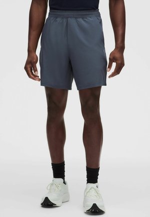 Modelo masculino con pantalones cortos deportivos gris oscuro, calcetines negros hasta el tobillo y zapatillas blancas, de pie frente a un fondo claro y liso.