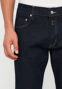 Jeans din denim de culoare albastru închis cu un design clasic, având un buzunar frontal, cusături galbene contrastante și un sistem de închidere cu buton metalic.