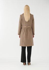 Brun uldblandingsfrakke med en defineret talje, guldknapsdetaljer og lange ærmer, parret med sorte bukser og højhælede sko.