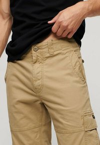 Pantaloni cargo beige con una texture liscia, dotati di molteplici tasche, chiusura con bottone e dettagli di cucitura visibili.