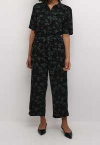 Combinaison noire à manches courtes avec motif de feuilles vertes, taille élastique et jambes larges effilées ; portée avec des chaussures noires à bout pointu.