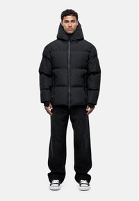 Black puffer jacket with a high collar and hood, featuring a zip front, oversized fit, and textured fabric. Worn with black jeans and sneakers.