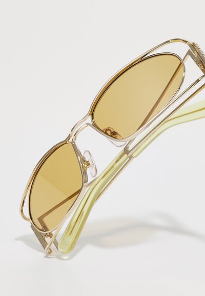 Sunglasses with an amber lens, transparent gold frame, and green temples. Features a sleek design with a unique shape and subtle accents.