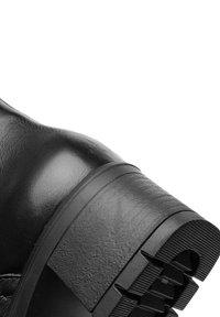 Botte en cuir noir avec un embout rond et une épaisse semelle en caoutchouc texturé. Le talon est épais avec des détails striés pour une meilleure adhérence.
