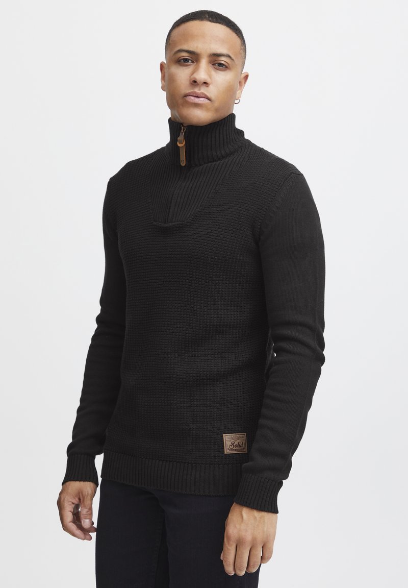 Black knit sweater with a high collar and half zipper. Features ribbed cuffs and hem, textured body, and a small logo patch at the waist.