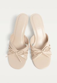 Beige keinokangas liukuva sandaali, jossa on litteä pohja, kahdella kapealla hihnalla ja koristeellinen sulka varpaiden alueella.