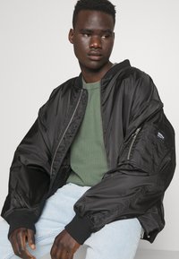 Giubbotto bomber nero in tessuto lucido con polsini a costine, dotato di zip frontale e tasche laterali. Abbinato a un maglione verde strutturato.