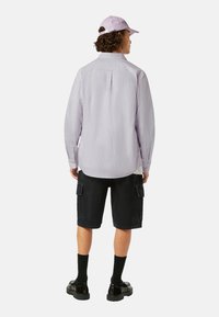 Chemise lilas à manches longues, shorts cargo noirs et chaussures noires avec des chaussettes. Couture arrière visible sur la chemise, casquette plate de couleur lilas assortie.