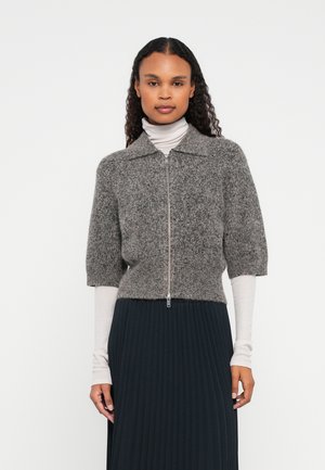 Žena v šedej zipsovej pletenej kardigane cez béžový rolák a čiernu plisovanú sukňu, stojaca pred bielym pozadím.