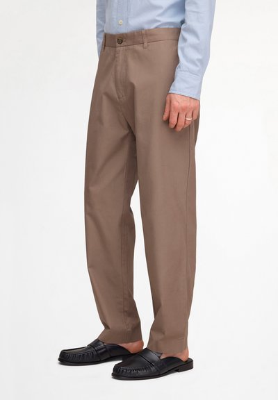 Homme portant un pantalon droit marron, une chemise bleu clair boutonnée et des mocassins noirs sans lacets, debout devant un fond blanc.