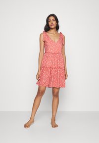 Rode bloemenjurk met korte, gestrikte bandjes, V-hals en gelaagde rok. De stof heeft een lichte textuur met kleine witte en roze bloemenpatronen.