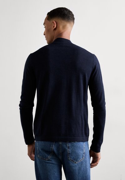 Pull long bleu marine en maille lisse avec un col rond, associé à un jean en denim bleu clair avec des poches arrière.