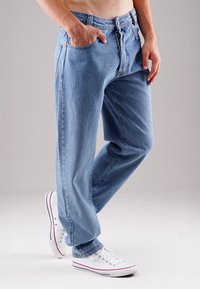 Hellblaue Denim-Jeans mit geradem Schnitt, ausgestattet mit einem Reißverschluss und mehreren Taschen. Getragen mit weißen Sneakers.
