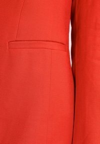 Röd texturerad tyg, skräddarsydd design, med en diskret ficka. Materialet visar ett fint vävt mönster med en slät yta.