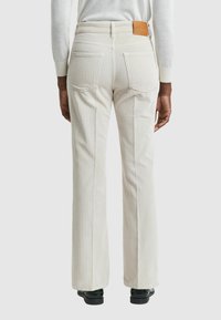 Pantalons en velours côtelé couleur crème avec jambes évasées, poches arrière et une étiquette en cuir marron au niveau de la taille. Tissu texturé avec des côtes verticales.