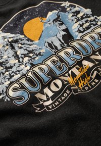 T-shirt en coton noir avec un imprimé vintage de montagnes, d'un cerf et d'une lune jaune. Le texte comprend "SUPERDRY" et "Mountain High" en couleurs vives.