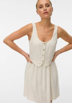 Kvinde iført ærmeløs, knaplukket beige kjole med tætsiddende krop og udvidet skørt, hænder hvilende på hofterne, neutral baggrund.