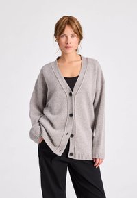 Lichtgrijze gebreide cardigan met een V-hals, lange mouwen, geribbelde manchetten en vier zwarte knopen aan de voorkant. Zachte, middelzware textuur.