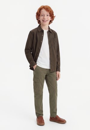Rothaariger Junge lächelt, trägt ein dunkelgrünes Hemd mit Knöpfen über einem weißen T-Shirt, olivfarbene Cargohosen und braune Slipper, steht mit den Händen in den Hosentaschen.