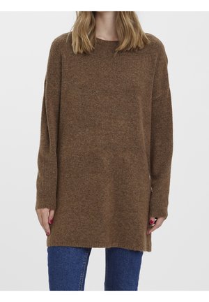 Kvinde iført en lang brun strikket sweater med rund hals og blå jeans, stående foran en ensfarvet lys baggrund.